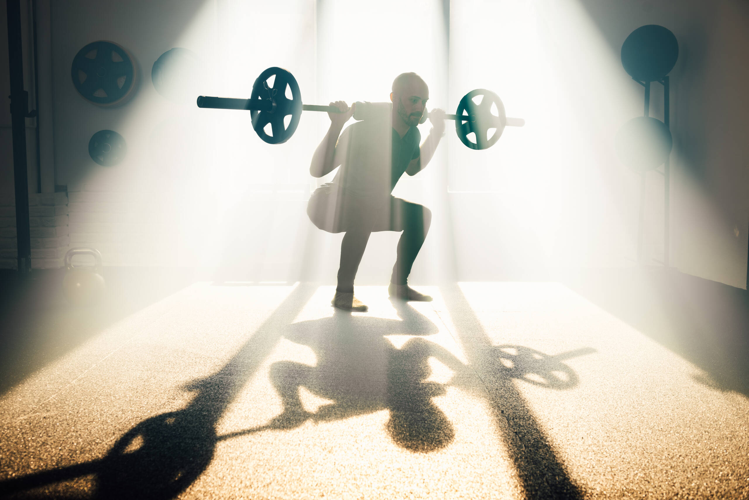 Strong man lifting barbell in gym in sunlight Side view of confident male athlete in sportswear looking down while standing on bent knees with barbell on shoulders in back lit sunbeam with shade in gym and exercising alone